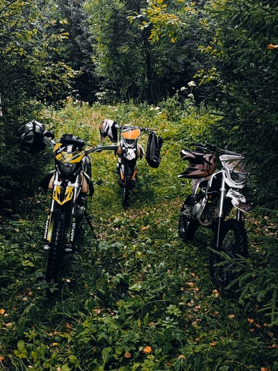 The image depicts three dirt bikes parked in a wooded area. The bikes are arranged in a triangular formation, with one in the foreground and two slightly behind and to the sides. The bike in the foreground is black and yellow, with the brand "MOTUL" visible on its front fender. Helmets are placed on the handlebars of each bike. The surrounding environment is dense with green foliage, including trees, bushes, and grass. There are also some fallen leaves scattered on the ground. The lighting suggests an overcast day or late afternoon. There are no people visible in the image. The scene appears to be a trail or off-road area within a forest.