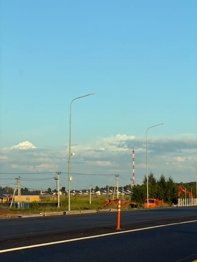 The image depicts a construction site on the outskirts of Kaipy, Russia, with a clear blue sky and scattered clouds. In the foreground, a freshly paved asphalt road with a white dividing line is visible. A bright orange traffic cone stands near the right edge of the road.

Further back, several modern houses with light-colored facades are lined up in a semi-rural setting, suggesting a developing residential area. Several utility poles with electrical wires crisscross the scene.

Dominating the middle ground are construction vehicles, including an orange truck and a red excavator, positioned near piles of dirt. A tall, red and white striped communication tower rises in the background. Green trees and shrubbery are interspersed throughout the landscape. The overall impression is one of ongoing infrastructure development in a newly developing area.