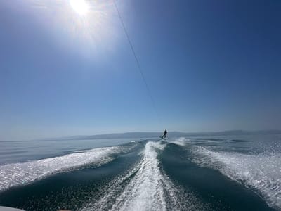 The image displays a wakeboarding event on the Sea of Galilee, near Tiberias, Israel, under a bright, clear blue sky. A single human figure, silhouetted against a prominent sun flare originating from the upper left quadrant, stands on a dark wakeboard. This individual is holding a thin tow rope that extends upwards and out of frame to the left, indicating attachment to a towing vessel. The water surface is dark blue, marked by a significant, turbulent white wake originating from the immediate foreground, created by the observer's boat, and extending towards the wakeboarder. Smaller sprays of water are visible around the wakeboarder's board. Distant, low-lying landforms are visible along the hazy horizon line. The scene is brightly illuminated with strong backlighting.
