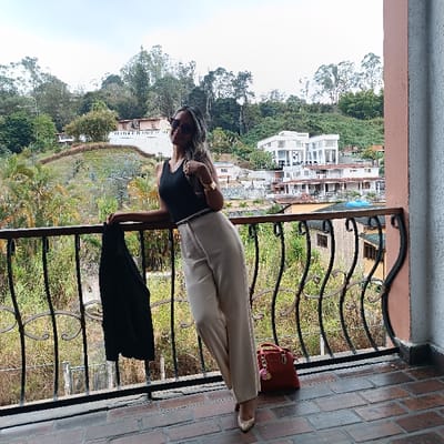 A woman in a black top and beige pants poses on a balcony with an intricate iron railing. She wears sunglasses, a watch, and a thin belt. She holds a black blazer draped over the railing. A red purse sits on the floor near her. The background shows a lush green landscape with buildings nestled amongst the trees under a partly cloudy sky. The balcony floor is brick.