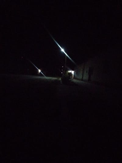 The image depicts a dark outdoor scene, likely at night, with limited illumination. A street light is prominently visible on the left side of the frame, casting a bright beam diagonally across the image. To the right, a low-rise building is partially illuminated by this light, revealing a doorway or entrance area with interior light. The ground in front of the building appears to be a pathway or road, with some subtle texture visible where the light falls. The overall environment is dark and appears to be an exterior space, possibly a street or courtyard area adjacent to a building. The location in Kargalar, Uzbekistan, is not visually discernible from the image itself.