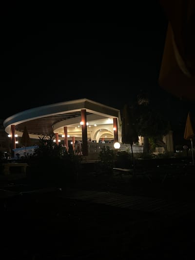 The image captures an outdoor seating area of a establishment at night, possibly a restaurant or bar, in Rhodes, Greece. A curved, covered structure with pillars illuminated by warm lights provides shelter and seating. Several people are visible in the background, some standing and others seated, suggesting a social gathering. Umbrellas, lounge chairs, and plants are scattered throughout the scene. The foreground is predominantly dark, with the light from the establishment casting some illumination on the paved ground. The overall ambiance is one of a relaxed evening setting.