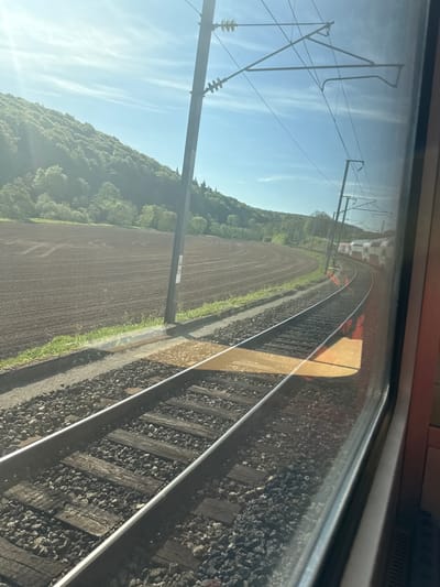 This media file captures a view from inside a train, looking out a window at a railway track curving into the distance. The perspective suggests the train is either moving or stationary on a bend.

In the foreground, two sets of railway tracks are visible, laid on a bed of dark grey ballast with wooden sleepers. Reflections on the train window are apparent, including what appears to be part of the train's interior structure and a warm, golden-yellow reflection stretching across the tracks and ballast.

Beyond the tracks to the left, a vast, freshly tilled brown agricultural field extends outwards. Further in the background, a densely forested hill or mountain range rises, covered in lush green trees. Overhead, the blue sky is bright with scattered, wispy white clouds. Railway infrastructure, including a prominent power line support pole and overhead catenary lines, is visible against the sky. One wooden pole next to the field has a small white sign with text, which upon closer inspection appears to include "2020" and other indistinct numbers or symbols.

The scene depicts a sunny daytime, likely morning or late afternoon, given the angle of the light. The weather is clear and bright. This rural landscape, with its combination of agricultural land, forested hills, and modern railway infrastructure, is located in Essingen, Luxembourg.