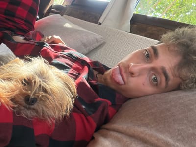 The image depicts a Caucasian male lying on a patterned beige cushion wearing a red and black plaid shirt. He has brown curly hair and is sticking his tongue out. A small, light brown dog is lying next to him. Outside, through a window, there is foliage visible. There is no information to support the assertion that the picture was taken in the city of Pripek, Bulgaria.
