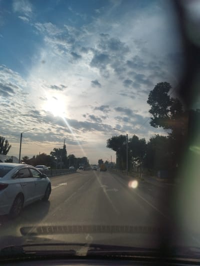 This is a wide-angle, eye-level, exterior shot taken from inside a vehicle, looking out at a road in Sokuluk, Kyrgyzstan. The sky is partially cloudy with the sun shining through, creating lens flare and streaks of light. The road ahead is paved with visible lane markings, including directional arrows. Several vehicles are present: a white sedan in the left lane, a yellow van further down the road, and other cars in the distance. Roadside features include trees, streetlights, and concrete barriers separating lanes. The environment appears to be an urban or suburban area.