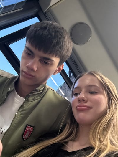 This is an upward-angled selfie featuring a young male and a young female, likely taken inside a vehicle during the daytime.

The male, positioned in the foreground and to the left, has short dark hair and prominent eyebrows, and is looking downwards with a neutral expression. He is wearing a dark green bomber jacket with light-colored stripes on the sleeves and a white t-shirt underneath. On his jacket, there is a red shield-shaped patch featuring a white castle or building design and the word "dorthe" (or similar) written in white text above it. A white tag with the letters "li" is partially visible below the jacket's zipper.

The female, with long blonde hair, is leaning her head on the male's shoulder or upper arm. She is looking towards the camera with a relaxed and slightly amused or content expression. She is wearing a dark top or jacket.

In the background, a clear blue sky is visible through a window, indicating it is daytime. Above the individuals, a round, gray speaker is mounted on the ceiling of the vehicle. The interior structure suggests a public transport setting like a bus, train, or tram. The overall scene depicts a casual, intimate moment of two individuals together, potentially traveling in Borisova Griva Massiv, Russia.