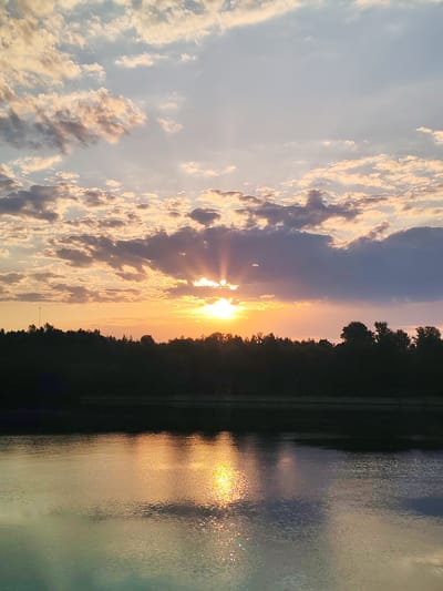 The image shows a sunset over a body of water, possibly a lake or river. In the foreground, the water reflects the colors of the sky and the setting sun. A line of trees is silhouetted against the bright horizon. The sky is filled with clouds of various shades of gray and orange, with the sun peeking through a gap in the clouds, casting rays of light. The location is possibly Ermolino, Russia, based on the query, although there is no specific indication within the image itself.