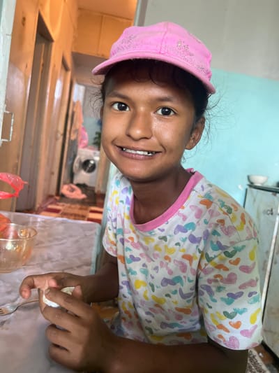 A young girl wearing a pink baseball cap and a t-shirt with a heart pattern is seated at a table, holding a piece of food. She has dark hair and is smiling, showing her teeth. She is holding a small, white object in her hands, possibly food she is preparing or eating. A fork is visible on the table next to her. In the background, there is a hallway with doors, a washing machine, and a white bowl on a shelf. The scene appears to be indoors, in what looks like a home or a communal living space.