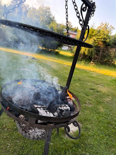 In Mordarka, Poland, a metal barbecue pit sits outdoors on a grassy lawn. Coals and burning wood are visible within the pit, generating a plume of smoke that rises and drifts. A metal grill grate is suspended above the fire, attached by chains to a metal frame. The background shows trees and a house, suggesting a residential area. The sun is setting, casting a warm glow on the scene. No people are present.