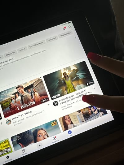 A close-up, slightly angled shot captures a person interacting with a black tablet device in a dimly lit indoor setting. Two fingers, adorned with red nail polish, are prominent in the foreground, one resting on the device's bezel and the other hovering over the screen.

The tablet's screen displays a bright interface, likely a video streaming application such as YouTube, showing a feed of recommended videos. The top left corner indicates a full battery at "100%". Visible navigational tabs and categories include "Yeni öneriler" (New recommendations), "İzlenenler" (Watched), "Son yüklenenler" (Last uploads), "Turizm" (Tourism), and "Sitcom'lar" (Sitcoms), all in Turkish. A search icon and a notification bell with '8' new notifications are also present.

Two prominent video thumbnails are clearly visible. The top one features a woman in a yellow dress within a dramatic, dark setting, identified as "Ariana Grande - hate that i made you love me (official m...)". It's 5:01 long, was uploaded 17 hours ago, and has 3.5 million views. Below it, another video thumbnail shows a group of young people, titled "Daha 17 | 1. Bölüm" (More 17 | 1. Episode), with a duration of 2:21:53, uploaded 1 day ago, and accumulating 4.4 million views. A partially visible third thumbnail shows a man and woman, featuring the title "KARA SEVDA" and a "1080p FULL HD" badge.

Faint German text "en Aufwand und übergib nur relevante Gespräche ans Team. o.ai" is visible at the bottom left, suggesting either the device's locale or the user's language preferences. The tablet also shows links for "Ana Sayfa" (Homepage), "Shorts", "Abonelikler" (Subscriptions), and "Siz" (You).