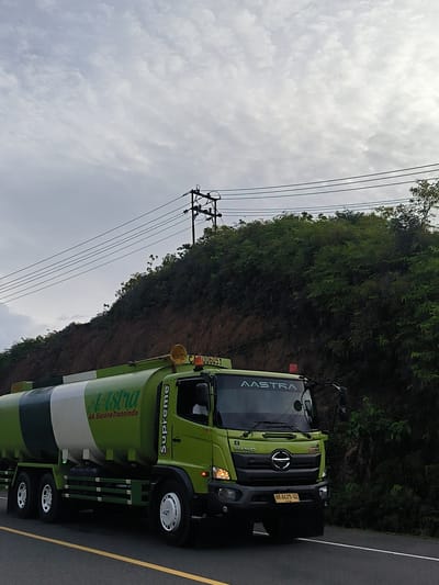 A bright green and white tanker truck with "AAstra" and "AA. Sarana Transindo" written on its side drives on a paved road. The truck has the word "Supreme" written vertically on its side door. It also has the Hino logo on the front. The truck is on the right side of the road, moving away from the camera. The truck has a license plate that reads "BW 8425 GI". In the background, there is a steep, vegetated hill with power lines running across it. The sky is overcast. The scene appears to be in Keudeunga, Indonesia.