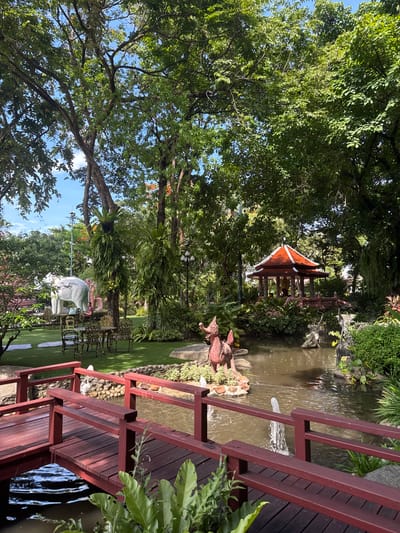 The image depicts a serene outdoor scene, likely a garden or park setting in Samrong Nuea Subdistrict Municipality, Thailand. A pond or small lake is visible with a decorative gazebo or pavilion situated on its bank, surrounded by lush greenery and trees. A wooden bridge with red railings crosses part of the water feature. Manicured lawns and ornamental sculptures are visible. The sky is partially visible through the trees, indicating a bright day.