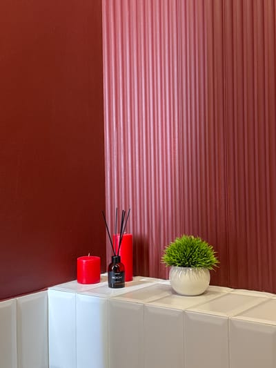 The media file displays a close-up, vertically oriented shot of a decorative arrangement on a surface. The background features a textured red wall with horizontal lines. To the right, a white tiled surface with a geometric pattern is visible.
On the tiled surface, a small, round, white pot contains a bright green, spiky artificial plant. Next to the plant, a reed diffuser with a dark glass bottle and black reeds is placed. The bottle has a label with the text "MEROMI" and "COTTON" printed on it. Beside the diffuser are two red cylindrical candles.
The lighting appears to be indoor, with the light source illuminating the objects from the right. The overall scene suggests a decorative corner in a room. There are no people, and no specific time of day or weather conditions can be inferred. The location context provided is Патрушева, Russia, which is not visually evident from the image itself.