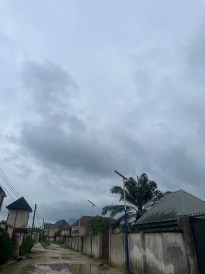 An overcast sky looms above a semi-urban street in Elimgbu, Nigeria. The street is unpaved and appears to have recently experienced rainfall, with several large puddles reflecting the gray sky. On either side of the street are residential buildings with concrete walls and various roofing materials, including corrugated metal and asphalt shingles. Some buildings are simple, while others are more elaborate with decorative elements. Vegetation, including palm trees and bushes, is visible throughout the scene. Electrical poles with wires run along the left side of the street, and streetlights are present on poles along the right. A sign is partially visible on the left, near a fence, with some text that appears to be religious in nature. The overall impression is one of a developing residential area under a cloudy sky.