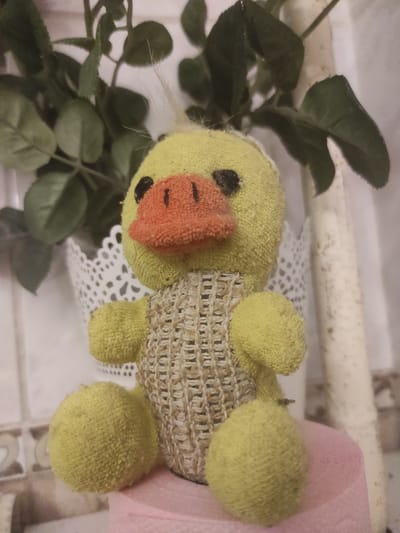 A close-up, low-angle shot depicts a yellow plush duck toy sitting on a pink surface, with a potted plant and a decorative white lace planter in the background. The duck is facing forward, with a bright orange beak, black eyes, and a textured brown weave across its chest. Its body is made of a soft, yellow terrycloth material. The plant has dark green leaves and appears to be artificial. The white lace planter has an intricate, delicate pattern. The background is blurred, showing hints of a tiled wall and a tall, cylindrical object that is off-white in color. The lighting is dim, creating a soft focus effect. The scene is indoors, likely in a home environment. The location is Yelizavetinskaya, Russia.