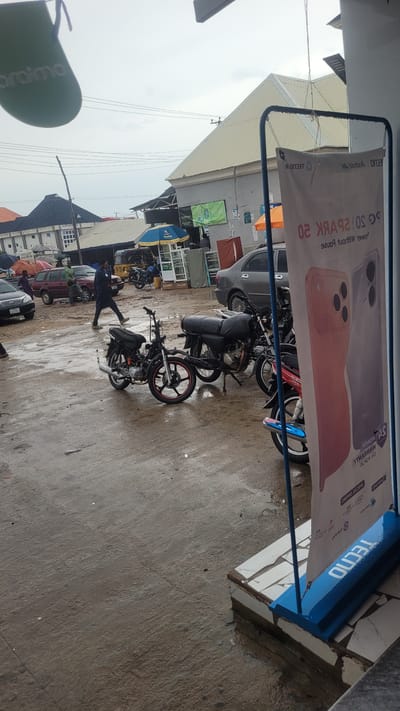 This is a rotated, eye-level, outdoor photograph taken during the day. The scene depicts a street or outdoor market area with several motorcycles parked on a wet, paved surface. A large banner hangs from a blue metal frame, displaying promotional text for "TECNO" and an advertisement for "SPARK 20" phones. The banner features images of two mobile phones.
In the foreground, a person wearing dark clothing and white shoes is in motion, appearing to walk or jump. They are positioned near a motorcycle. Several other motorcycles are visible in the background, along with parked cars and what appear to be market stalls or shops. The ground is wet, suggesting it may have recently rained or that the area is prone to puddles.
The sky is overcast and appears bright, indicating daylight. The overall impression is of a busy, functional outdoor space, possibly in a tropical or subtropical region, given the presence of motorcycles and what might be market activity. The visible text on the banner is primarily promotional, advertising mobile phone models and brands. Other smaller signs and logos are present but less legible.