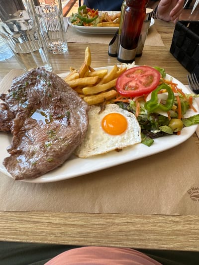 The image captures a meal served on a white, oblong plate, placed on a light-brown wooden table. The meal consists of a piece of steak, cooked but not heavily browned, topped with a glistening sauce or oil; fried potatoes, mostly golden in color; a fried egg with a bright orange yolk and a slightly browned white; and a small salad. The salad includes lettuce, shredded carrots, sliced green bell pepper, sliced tomato, and olives. The sauce in a bottle visible on the table suggests a casual dining atmosphere. There's a metal container to the left of the plate holding the sauce bottle and some visible drinking glasses. A person's hand can be partially seen at the top-left of the image. Another person's clothing is present on the right side. The image has a stamp of a "Gurb Nature" logo which suggests the location might be in Gurb, Spain.
