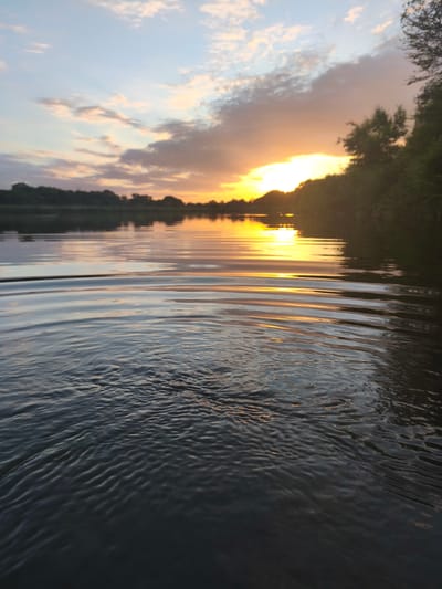 The image shows a body of water, possibly a river or lake, with a sunset in the background. The sky is filled with clouds and the sun is setting on the horizon, casting a warm glow on the water. The water's surface is rippled, reflecting the colors of the sunset. Lush green trees and vegetation line the far bank of the water. The foreground of the image is dominated by the water's surface, with a close-up view of the ripples. There are no people or any visible man-made structures in the image.