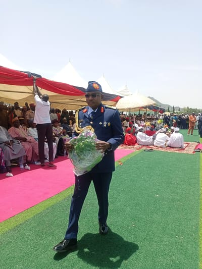 A man in a dark blue military uniform stands in the foreground, holding a clear-wrapped floral arrangement. He wears a matching cap and sunglasses. Behind him is a gathering of people under tent-like structures, with some seated on a pink carpet and others on a woven mat. A man is seen adjusting the fabric of the tent structure. The scene takes place outdoors on a green surface, possibly artificial turf, with trees and hills visible in the distance. Based on the prompt instructions, the location is in Dilpurtu, Nigeria.