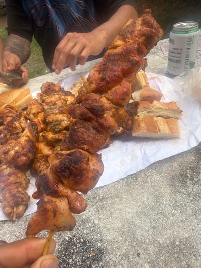 The image depicts a close-up view of a table laden with food, likely outdoors given the presence of a concrete surface and greenery in the background. The primary focus is on several skewers of grilled chicken, with one skewer prominently held in a person's hand in the foreground. The chicken appears well-cooked, with a slightly charred exterior. Adjacent to the chicken skewers are slices of bread, some appearing to be baguette. A can of beverage, possibly beer, is visible to the right of the food. The scene suggests a casual gathering or picnic. The reference to Campezo / Kanpezu, Spain, is contextual information not directly discernible from the visual elements of the image itself.
