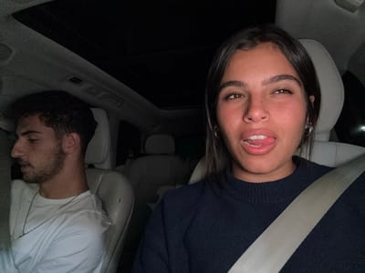 The image displays the interior of a vehicle at night, featuring two individuals. In the foreground, on the right, a female with dark hair wears a dark blue sweater and a light-colored seatbelt diagonally across her chest. Her face is illuminated, and she is captured with her tongue slightly extended, looking towards the right side of the frame. She has silver hoop earrings visible. To her left, in the midground, a male with dark, curly hair and facial stubble is seated, wearing a white t-shirt and a silver chain necklace. He is seen in profile, looking downwards with his eyes appearing closed or resting. The vehicle interior features light-colored seats and a visible sunroof mechanism on the ceiling. The background outside the vehicle is completely dark, indicative of nighttime. The specific location within Lawrence Township, United States, is not discernible from the visual evidence provided in the image.