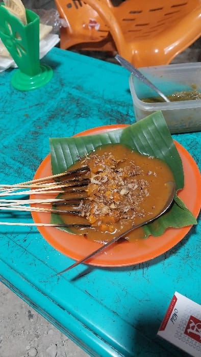 The scene is set in Alue Mudem, Indonesia, depicting a close-up view of a meal on a weathered, teal-colored table surface. The primary subject is an orange plastic plate containing multiple skewers of satay, arranged on a banana leaf. The satay is heavily coated in a thick, brown peanut sauce and topped with crumbled, fried shallots. A silver spoon is partially submerged in the sauce on the plate. To the left, a green plastic utensil holder, designed with cut-out floral patterns, contains several wooden toothpicks or small skewers. In the upper right quadrant, a clear plastic container holds a green, liquid sauce, possibly sambal, with another silver spoon resting inside. A segment of an orange plastic chair is visible in the blurred background. In the lower right corner, a white and red pack of "LA Mild" cigarettes is visible. The ground below the table appears to be concrete or gravel. No individuals are present in the image.