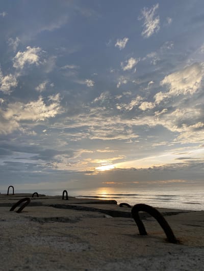 An eye-level shot captures a concrete structure with rusted metal loops protruding from it. The structure runs along the coastline, with the ocean visible in the background. Waves gently lap the shore under a sky filled with dramatic clouds. The sun is setting, casting a golden glow on the water and clouds. The scene appears to be a public space, possibly a promenade or a breakwater, offering a view of the sea and sky. The location is described as Bateetimoh, Indonesia.