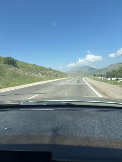 The image captures a view from inside a vehicle, looking out through the windshield. The perspective is slightly tilted, suggesting the vehicle is on a road. The scene depicts a mountain landscape with a clear blue sky and scattered white clouds. A paved road with white lane markings runs through the center of the frame, disappearing into the distance. To the left of the road, a lush green mountainside slopes upwards, dotted with trees. A white guardrail with reflective elements is visible along the edge of the road, indicating it's a mountain pass.
The right side of the image shows the interior of the vehicle, specifically a textured, dark-colored dashboard. A subtle reflection of a circular object, possibly a logo or emblem, is visible on the road surface. The time of day appears to be daytime, judging by the bright sunlight and clear sky. The weather is sunny and pleasant, conducive for travel. The overall impression is that of a scenic drive through a mountainous region. There is no visible text in the image.