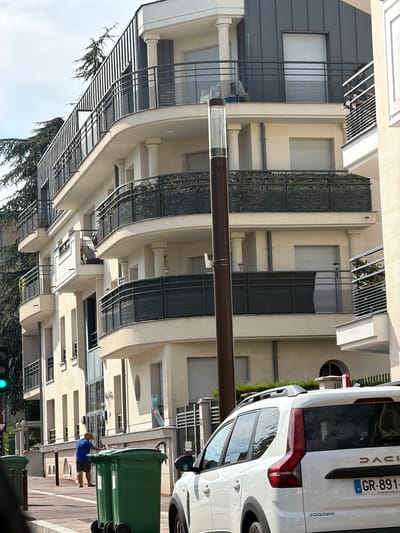 In Châtenay-Malabry, France, a person in a blue t-shirt and grey shorts is seen leaning against a stack of large green trash bins on a street. The scene appears to be outdoors, with a building featuring cream-colored facade and dark metal balconies or sunshades in the background. A white Dacia Jogger vehicle is parked on the right side of the frame, with its rear visible. The license plate reads "GR-891-NT". Above the car, the word "DACIA" is visible. The building has multiple floors with windows and architectural details like pillars and curved balconies. Trees with dark green foliage are visible in the lower left corner of the image, suggesting it might be daytime. A traffic light showing a green signal is also visible in the lower left. The overall impression is an urban street scene on a clear day.