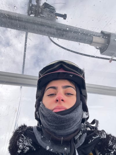 A young woman is depicted in a close-up, head-and-shoulders view from inside a ski lift enclosure in Valtournenche, Italy. She looks directly at the camera with a neutral expression, wearing a black ski helmet with dark goggles pushed onto the forehead, and a grey neck gaiter covering her lower face, leaving only her eyes and forehead exposed. Her nose appears reddish. She is dressed in a dark winter jacket featuring a faux-fur collar, which is partially covered in visible snowflakes or ice particles. The immediate environment is characterized by the transparent plexiglass or plastic roof and side panel of the ski lift, which is coated with numerous water droplets. Through this clear material, metallic structural components of the ski lift apparatus are visible against an overcast sky.
