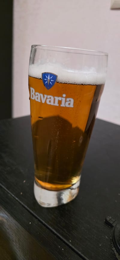 A close-up, eye-level shot shows a glass of Bavaria beer. The glass has the brand logo, a blue shield with a white starburst, and the word "Bavaria" written in white letters. The beer itself is a clear amber color with a frothy head and small bubbles visible throughout. The glass is sitting on a dark, textured surface, possibly a wooden table. The background is a soft, out-of-focus light pink wall. There are no people visible in the image, and the scene appears to be indoors. The image does not contain information to indicate its location in Gori, Georgia.