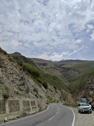 The image captures a scenic mountain road in India, likely in a mountainous region given the terrain and vegetation. The road curves through a valley with steep, rocky slopes on one side and verdant, green mountains in the distance. The sky is partly cloudy with fluffy white clouds against a pale blue backdrop.
On the left side of the road, a retaining wall constructed with stones and concrete supports the rocky embankment, with some sparse vegetation growing from it. Power lines are visible traversing the upper slopes.
On the right side of the road, a white SUV is parked with its doors open, and two individuals are standing near it. Another person can be seen walking on the road further ahead, near a protective barrier along the edge. A small, rustic structure with a blue roof is also visible behind the parked car. The road itself is asphalt with white lane markings. The overall scene suggests a travel or tourist location in a natural, mountainous environment.