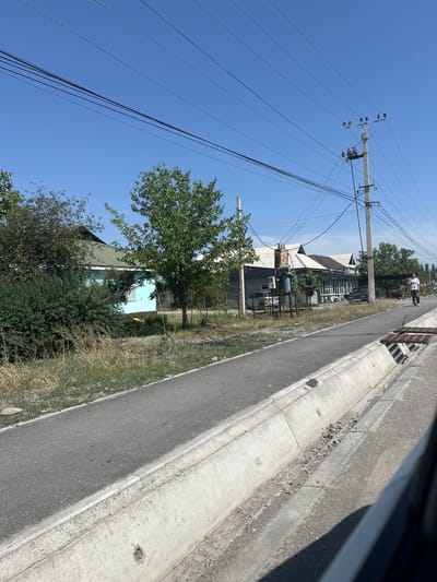 The image depicts a street scene in the city of Ынтымак, Kyrgyzstan, under a clear blue sky. A paved road runs diagonally across the frame, bordered by a concrete drainage ditch on the right and a grassy area with trees and bushes on the left. In the distance, several buildings are visible, including a light blue house with a green roof, and other structures with corrugated metal roofs, one of which appears to be a business with outdoor seating. A person in a white t-shirt and dark pants is walking away from the camera on the right side of the road. Utility poles with numerous power lines are prominent features of the landscape, stretching across the sky. The overall impression is of a residential or mixed-use area with typical urban infrastructure.