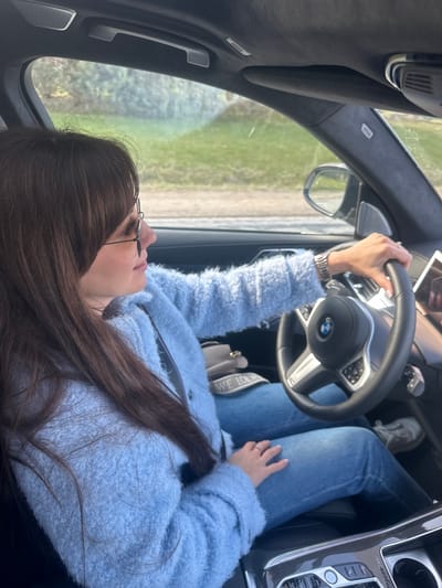 The image captures a top-down view inside a modern car, focusing on a person in the driver's or front passenger's seat. A female with long dark brown hair, wearing glasses, a light blue fuzzy jacket, and blue jeans, is visible from the chest down to her knees. Her left hand is gripping the steering wheel, which features a BMW logo. A watch is visible on her left wrist, and a ring is on her right hand, which rests on her thigh. She is also wearing light-colored sneakers.
The car's interior is dark, with visible details including a dashboard, a center console with carbon fiber-like accents, and various control buttons. Outside the windows, a blurred green and brown landscape suggests the car is moving through a natural or rural setting, consistent with the location context of Jaunsaurieši, Latvia. The time of day appears to be daytime, with ample natural light.
Visible text includes the "BMW" logo on the steering wheel, "AIRBAG" on the A-pillar, and several labels on the center console buttons: "OFF," "START STOP ENGINE," "SPORT," "COMFORT," "ECO PRO," and "AUTO H." A partial brand name "WE LOEW" (likely "LOEWE") is visible on the strap of a brown bag resting near her lap.