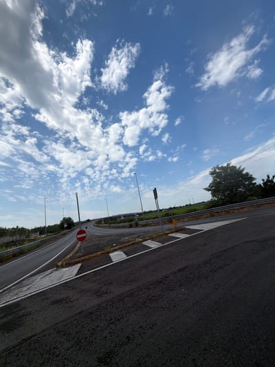 The image shows a road with a bright blue sky and scattered white clouds. The road appears to be asphalt and has white markings. There are streetlights along the side of the road, and a red and white circular sign is visible. In the background, there are trees and power lines, suggesting a rural or semi-rural setting. The overall impression is of a clear day, likely daytime, given the brightness of the sky. The image is taken from a slightly elevated perspective, looking down the road. The perspective is tilted, suggesting the photo was taken from a moving vehicle or handheld while the photographer was in motion. There is no discernible activity or event taking place, and no people are visible. The location context is Novara, Italy.