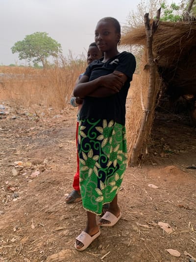 This image depicts two individuals in Tabu, Nigeria, within a dry, open outdoor environment. A female adolescent stands centrally, facing the camera with a faint smile, her arms crossed over her chest. She wears a black t-shirt and a long, patterned skirt featuring green, blue, and light green floral designs, along with light pink slide sandals. Behind her, partially obscured, is a younger child wearing a red garment and dark sandals, looking towards the left. To the immediate right of the main subject, a simple structure constructed from dry thatch or straw and wooden poles is visible. The ground consists of dry, reddish-brown earth with scattered debris and dry vegetation. In the background, there is an expanse of dry, straw-like plants, and a single large, green-leafed tree stands against a hazy, light-colored sky. Faint power lines are discernable in the upper left background.