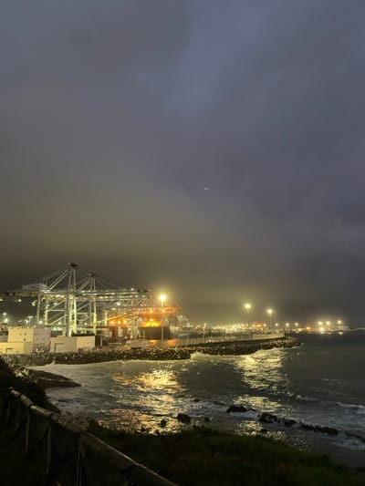 This forensic description details an illuminated port scene at night, located in Ksar El Majaz, Morocco, specifically depicting the Tanger Med port. The dark, overcast sky dominates the upper two-thirds of the image, showing faint traces of light penetration and possibly a faint streak from an aerial object in the mid-upper right. In the lower left foreground, a dark, indistinct grassy or rocky embankment is visible, partially obscured by a dark wooden fence or railing running horizontally.
The central focus is a large industrial container port. Multiple large white gantry cranes, equipped with red accents near their peaks, stand prominently. These cranes, along with several tall floodlights on the quay, brightly illuminate the scene, casting strong yellow and orange reflections on the agitated ocean surface. A large cargo ship, identifiable as a container vessel with visible containers on its deck, is docked at the quay beneath the gantry cranes.
To the right of the port, a long, curving breakwater constructed from large rocks extends into the dark water, protecting the harbor. This breakwater is also lined with several tall streetlights. The ocean exhibits moderate wave activity, with waves breaking against the shoreline and the breakwater, creating white foam. The artificial lights from the port and the breakwater intensely reflect off the water's surface, creating shimmering light paths. Numerous additional industrial lights are visible along the distant coastline. No individuals are discernible within the frame; however, the active lighting and visible cargo ship suggest ongoing port operations.
