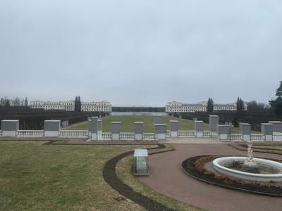 The image shows a symmetrical garden in Arkhangel'skoe, Russia, likely within the Arkhangelskoye Estate. In the background, two large, multi-story buildings with classical architectural details are visible on either side of a wide lawn that slopes down to meet the viewer's vantage point. The lawn is bordered by neatly trimmed hedges and pathways.
In the middle ground, a low wall with ornate balusters runs across the scene, punctuated by square pillars. In the foreground, there are several elements: On the left, a section of grass with a curved, dirt path containing small plants. Further right is a fountain with a tiered design.
The weather appears overcast with a grey sky. No people are visible in the scene. A glass case containing a sculpture or artifact is to the left of the fountain.