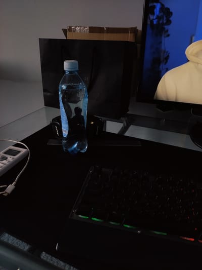 The image shows a desk setup with a keyboard, a bottle of water, and a computer monitor. The keyboard has illuminated keys, with some green and red lights visible. The bottle of water is clear and partially filled. In the background, there is a black bag and a cardboard box. The computer monitor displays a person wearing a light-colored hoodie. The overall scene is dimly lit, suggesting it might be nighttime or a room with low lighting. The location in Zhanadaur, Kazakhstan is not discernible from the image itself; this detail is provided as external information.