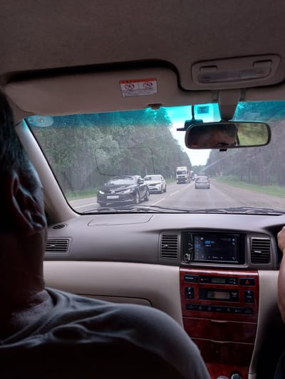 This media file captures a view from inside a car, looking out the windshield and at the dashboard. The scene appears to be a road trip on a highway, with a line of cars ahead, suggesting a traffic jam or a slow-moving journey. The surrounding environment is a forest, indicating a rural or semi-rural location, possibly in Russia as suggested by the OCR text.
The dashboard displays an audio system with a digital clock showing "17:03" and temperature reading "+28.9 C", suggesting it's late afternoon on a warm day. The weather appears overcast, as indicated by the diffused light and the slightly hazy view of the road and trees.
In the rearview mirror, a person's face is visible, likely the driver or a passenger. The interior of the car shows a sun visor with some warning labels, typical of car interiors. The dashboard itself has wood-grain trim, adding a touch of sophistication to the car's interior.
The overall impression is one of a mundane travel experience, perhaps a journey to or from a destination like Баюновские Ключи, Russia, characterized by the slow progress of traffic on a forest-lined road under a cloudy sky. There are no strong emotional cues visible, just the quiet observation of the journey. The visible text includes car controls, radio information, and warning labels.