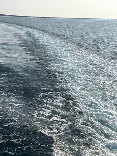 The image depicts a maritime scene off the coast of Kamariotissa, Greece. The foreground and midground are dominated by the turbulent wake of a moving vessel, characterized by intensely churned white foam and agitated dark blue-green water. This wake extends diagonally from the lower right corner towards the upper left. Beyond the immediate wake, the sea surface exhibits calmer ripples and a consistent deep blue hue. A faint, low-lying landmass, appearing as a light-toned strip, is visible along the distant horizon on the left side of the frame, indicating the coastline. The sky above is a uniform, pale, light-grey tone. No individuals, specific components of the vessel from which the image was captured, or other distinct objects are visible within the scene. The environment is open sea under daylight conditions.