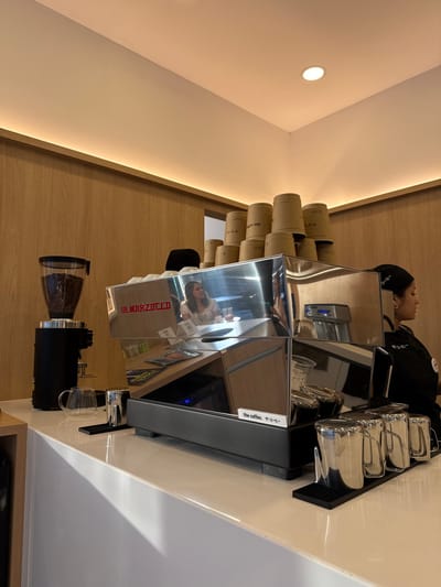 The image presents the interior of a coffee shop, identified by branding as "the coffee.", located in Torreón, Mexico. The central focus is a large, highly polished chrome La Marzocco espresso machine positioned on a glossy white countertop. The machine's reflective surface distinctly shows the reflection of a female patron seated at a table, wearing a light-colored top, with a beverage glass in front of her.

To the left of the espresso machine, a black coffee grinder with a transparent hopper full of dark roasted coffee beans is visible, alongside a small clear glass pitcher. To the right of the espresso machine, four stainless steel milk frothing pitchers are arranged on a black rubber mat. Directly above the espresso machine, approximately ten to twelve inverted brown paper cups, each printed with "the coffee. ザ・コーヒー", are stacked.

A female barista is partially visible on the far right of the frame, wearing a black uniform and cap, with her face turned to the right. The background features light brown wooden wall paneling. Illumination is provided by a continuous strip light integrated into the wall-ceiling junction and a single circular recessed light fixture in the white ceiling. The environment appears clean and modern.