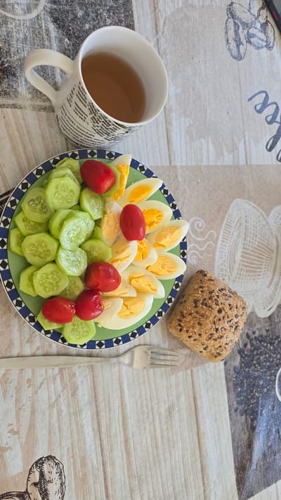 The image shows a breakfast setting on a patterned tablecloth. A plate with sliced boiled eggs, cherry tomatoes, and cucumber slices is centrally positioned. A whole grain roll is located above and to the right of the plate, and a fork is laid beside the plate. A cup of tea or broth is to the left. The location is Zirndorf, Germany, based on the visible texts on the mug.
