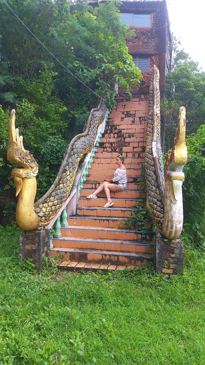 A woman poses on the staircase of Wat Tham Ta Pan temple in Thap Put, Thailand. She is wearing a striped top, denim shorts, and white sandals, and is positioned horizontally on the stairs, bending at the waist. The staircase is adorned with ornate dragon sculptures on either side. Lush greenery surrounds the staircase, with a building visible in the background. The staircase is made of orange bricks with blue accents.