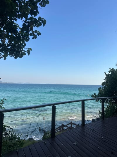 The image depicts a coastal view in Coolangatta, Australia, observed from a wooden deck. The ocean, a light turquoise color, is visible with faint city lines in the distance. A dark brown wooden deck with railings frames the right side of the image, while foliage is sparsely positioned on the left side and above. The sky is a clear blue, and the image has a slight tilt.