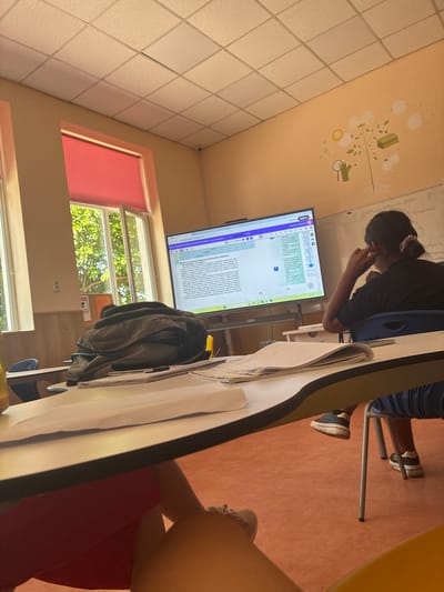 A classroom in Radievo, Bulgaria, features a large touchscreen monitor displaying text and educational graphics. A student, seen from the back, sits at a desk wearing a black shirt and has their hair in a bun. They are reaching up to adjust something near their ear while looking at the screen. In the foreground, a desk is covered with papers and a closed backpack. The room has large windows with red blinds, and the walls are painted a light orange with a cartoon-like mural of a plant and scientific tools. The ceiling is made of acoustic tiles. The floor is a warm-toned carpet.