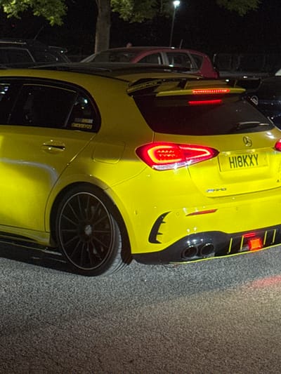 A yellow Mercedes-AMG A45 S car with registration number HI8KXY is parked on a street in the Borough of Spelthorne, United Kingdom. The rear taillights are illuminated. To the left is a car, visible only in part. The environment is urban with trees and street lights in the background. The surface on which the cars are parked is asphalt.