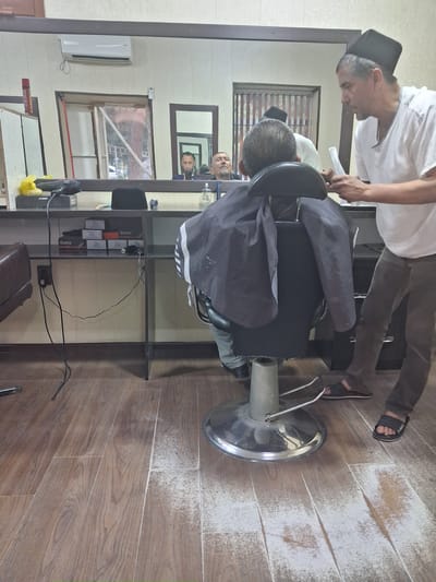 The image depicts a barber shop in Kahramon, Uzbekistan, with two individuals actively engaged in a haircut. A barber, wearing a white shirt and a black skullcap, stands over a seated customer who is covered with a dark cape. The barber is in the process of cutting the customer's hair with scissors and a comb. The customer, also wearing a skullcap, is looking down, seemingly focused on the haircut.
The setting appears to be a modest salon, evidenced by the presence of a professional barber chair, a mirror reflecting the scene, and shelves stocked with what appear to be grooming products. In the background, a large window shows a glimpse of the outdoors, suggesting it's daytime, though no specific weather conditions are discernible. An air conditioning unit is mounted on the wall. The flooring is made of wooden planks, some of which show signs of wear.
The interaction between the barber and the customer seems professional and routine. The overall atmosphere is that of a typical barber shop service. No explicit text is clearly legible in the image, other than possibly some branding on products on the shelves, which are too small to read.