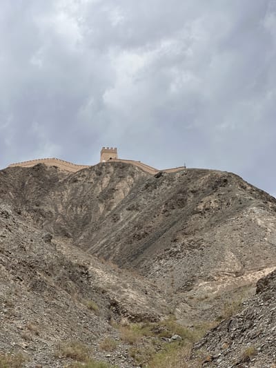 The image shows a section of the Great Wall of China, specifically the Jiayuguan Pass, an important landmark located in Jiayuguan City, Gansu Province, China. The Great Wall snakes along the crest of a steep, rocky mountain, its crenelated battlements and a watchtower visible against a cloudy sky. The terrain is rugged and arid, with sparse vegetation clinging to the scree-covered slopes. The watchtower is a prominent feature, built with tan-colored bricks, and appears to be well-preserved. There are several figures, appearing to be tourists, walking along the wall and near the watchtower, suggesting the area is accessible to visitors. The sky is overcast, casting a diffused light over the scene, and the overall impression is one of historical grandeur set against a harsh, natural landscape.