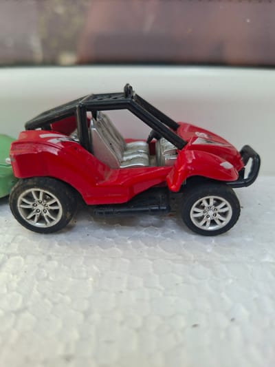 A red toy dune buggy is positioned on a white textured surface. The dune buggy has a black roll cage, silver seats, and silver rims on its tires. A portion of a green toy is visible to the left of the dune buggy. The background consists of a blurry surface with muted colors. The location in Gorkovskiy, Russia, is not discernible from the image.
